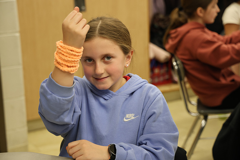 A fifth grade girl smiles and holds up her arm that has a peach colored bracelet that she crocheted.