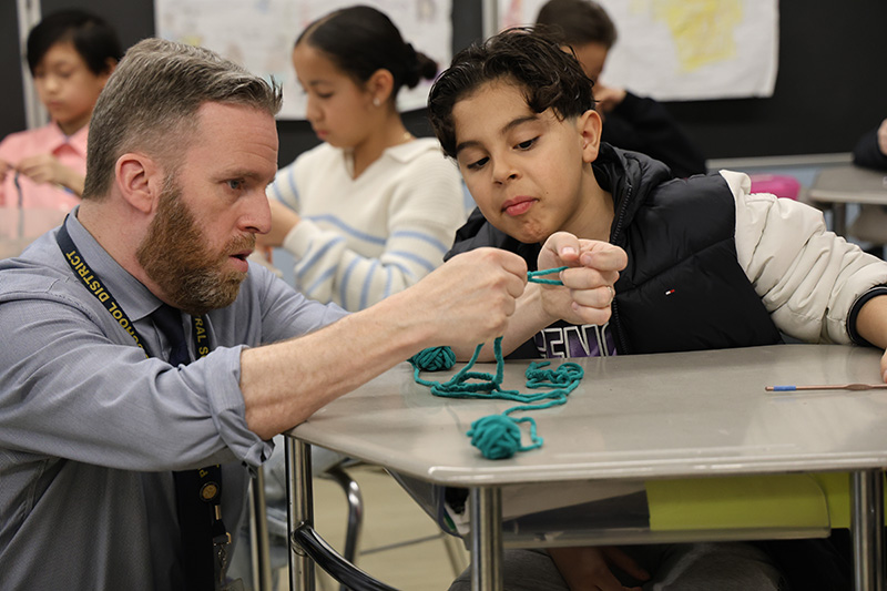 A boy sits at a desk and leans in while his teacher squats down and shows him how to crochet.