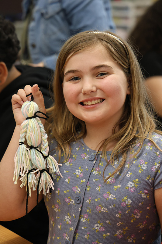 A girl with long blonde hair smiles an dholds up a little doll made of yarn.