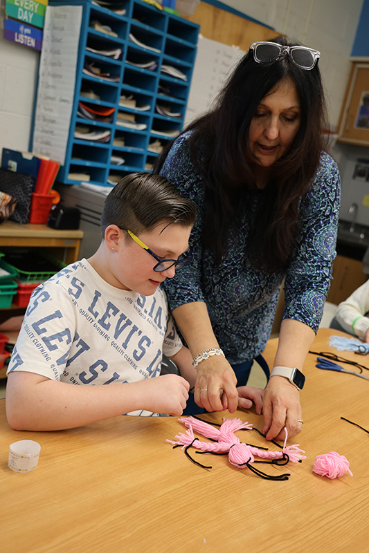 A woman with long dark hair leans over a student to help him make a doll made of yarn.
