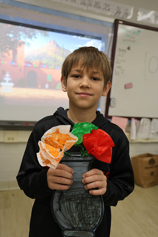A boy smiles as he holds up a paper vase with three paper flowers in it.