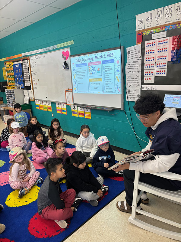 A high school kid wearing a football jersey reads a book to a class of elementary students.