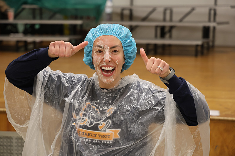 A woman wearing a plastic cape and a shower cap smiles broadly and gives two thumbs up with whipped cream on her face.