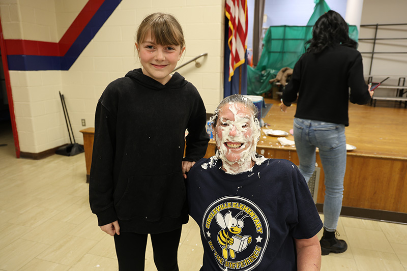An adult smiles with whipped cream all over her face. An elementary students stands next to her also smiling.