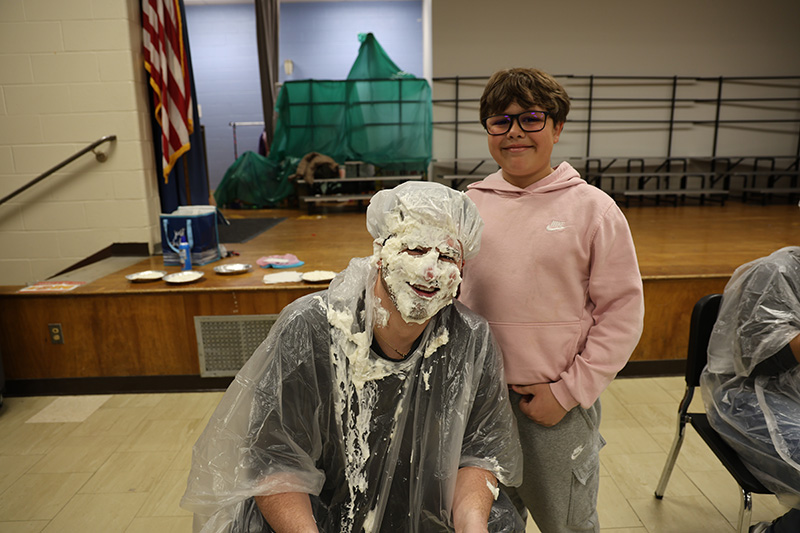 An adult smiles with whipped cream all over his face. An elementary student stands next to him also smiling.