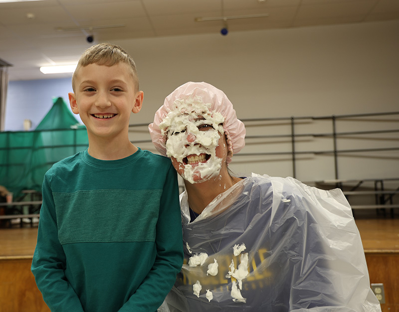 An adult smiles with whipped cream all over her face. An elementary students stands next to her also smiling.