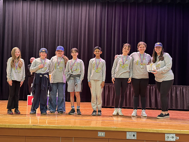 A group of seven middle school students and an adult stand on a stage with medals around their necks.