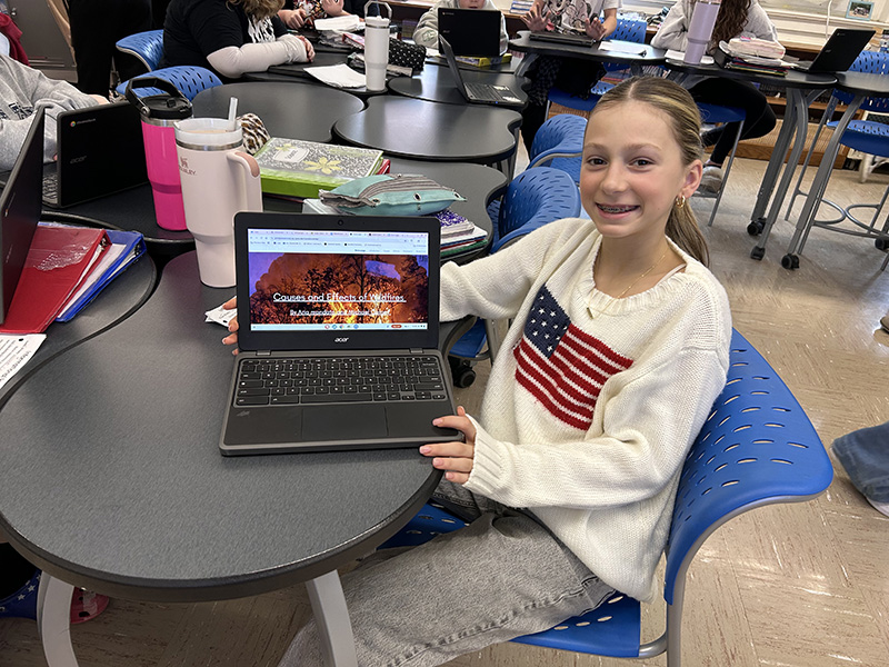 A middle school girl with long blonde hair smiles as she shows her ChromeBook with a website she created.