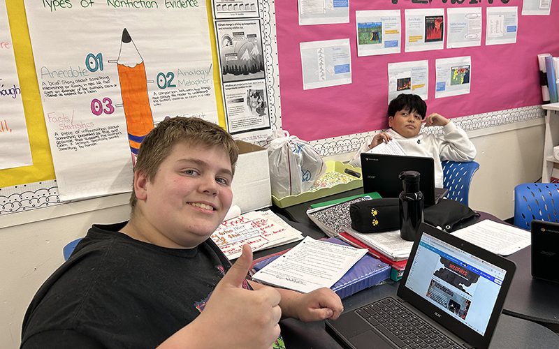 Two middle school boys sit at desks. One is giving a thumbs up showing the website he created on his Chromebook.