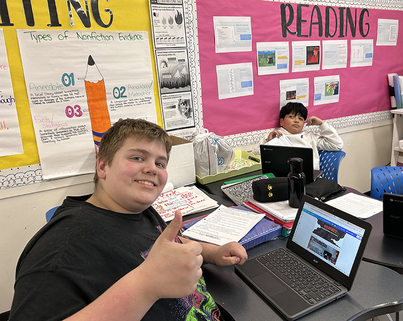 Two middle school boys sit at desks. One is giving a thumbs up showing the website he created on his Chromebook.