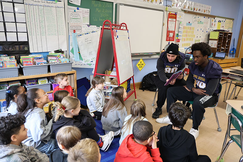 Two high school kids wearing football jerseys read a book to a class of younger elementary students.