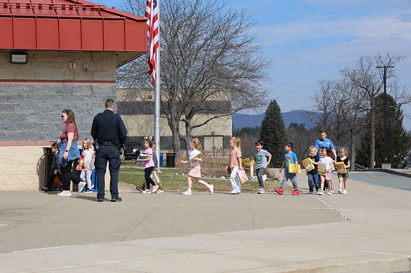 A line of kindergarten kids in a single file line walk to a big school.