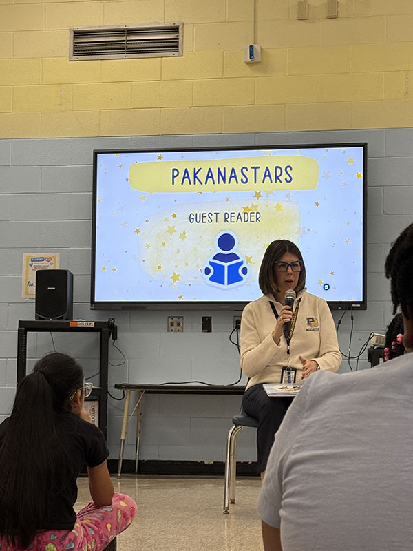 A woman sits in front of a group of kids and reads to them. Behind her is a screen that says Pakanastars.