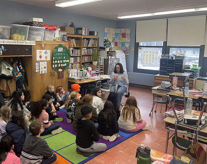 A woman sits in front of a group of children and reads to them.