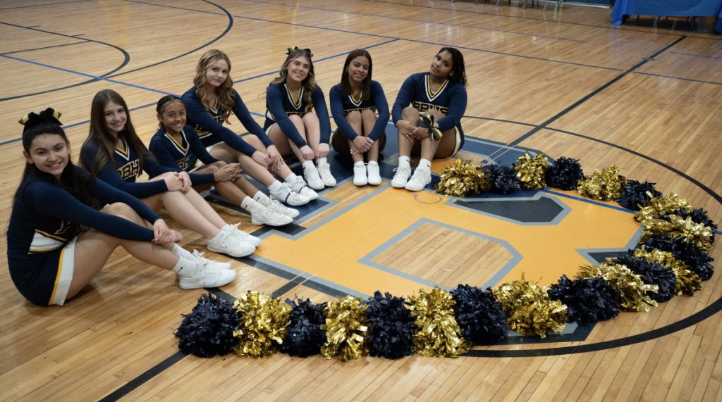 A group of cheerleaders in blue and gold sit around a circle at center court of  a basketball court. Their pompoms are lined up on the circle too.