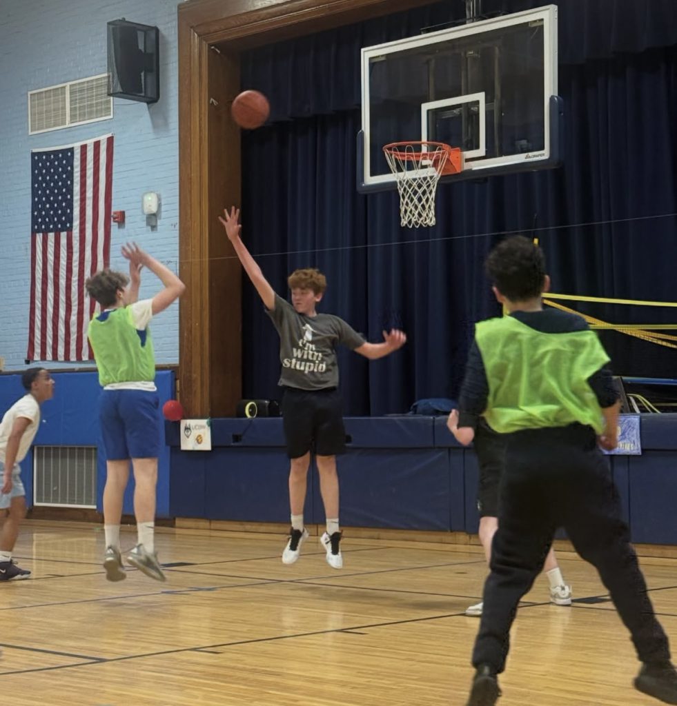 A middle school kid in a green pinny takes a basketball shot while another kid is jumping up to guard him.