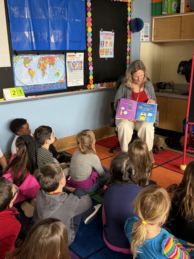 A woman sits on a chair in front of a group of kids and reads to them.