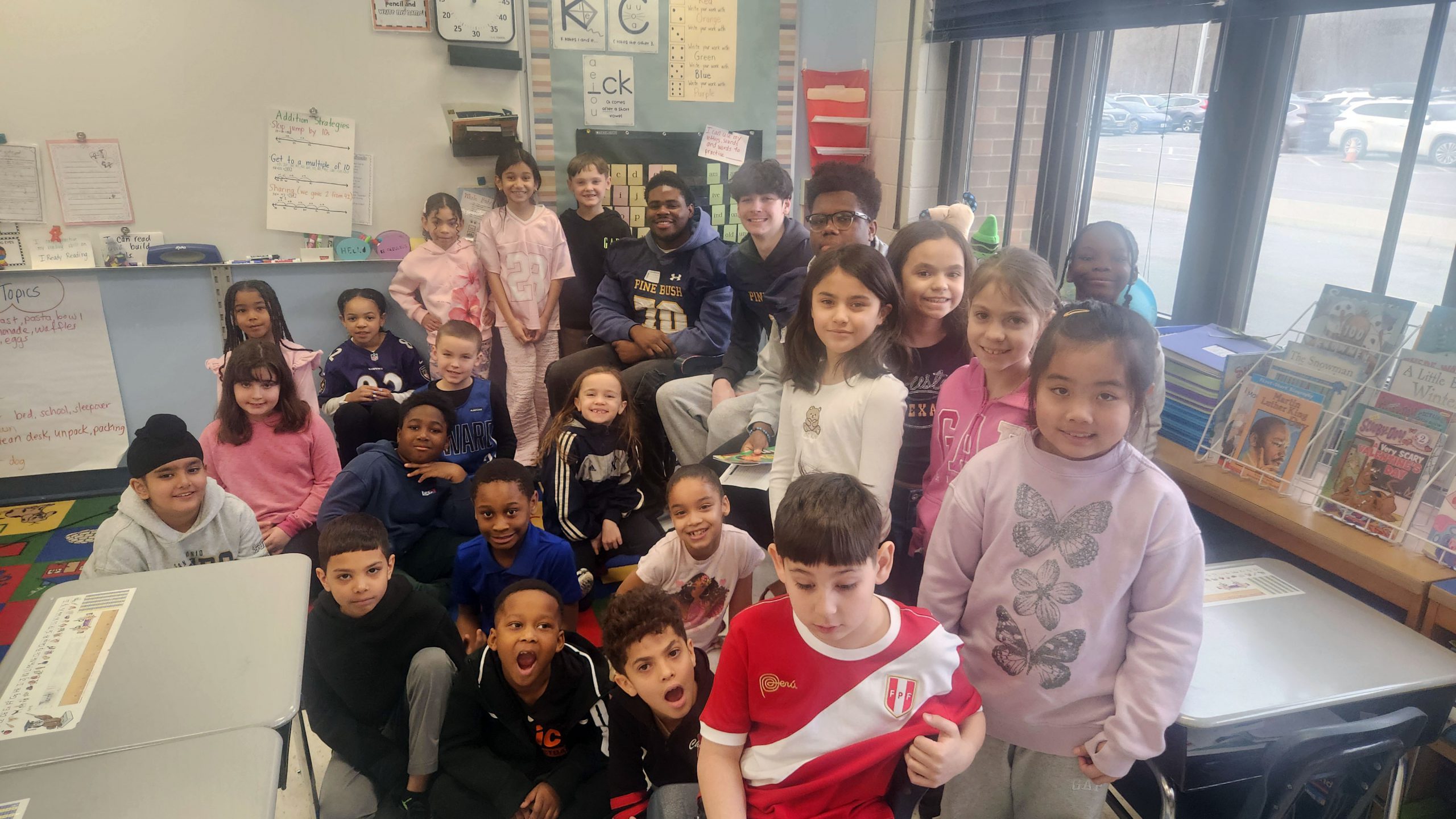 Three high school students dressed in football jerseys stand with a group of second grade students in a classroom.