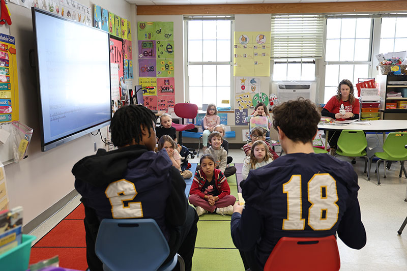 Facing a class of younger elementary students, two high school students wearing football jerseys read from a book.