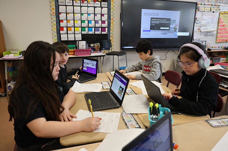 A group of four third-grade kids sit at desks working with chrome books, posters, crayons.