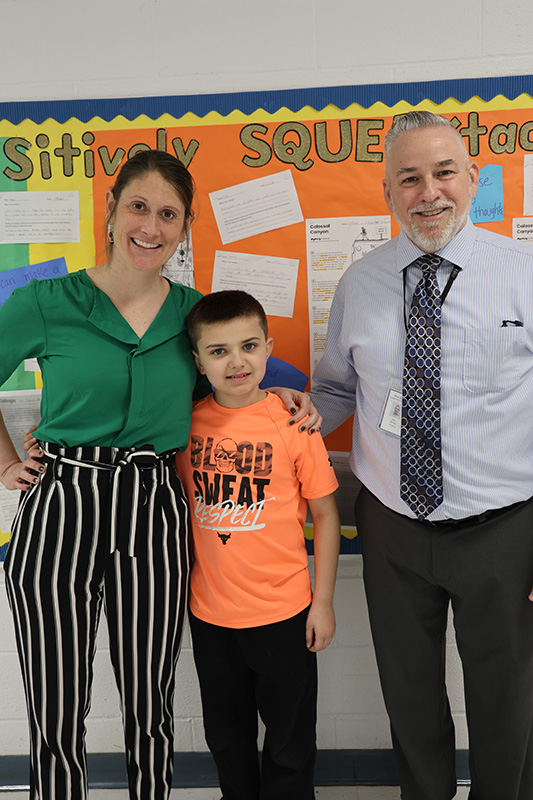 A woman in green shirt and black and white pants stands smiling with her arm around a boy wearing an orange shirt. Next to the boy is a man also smiling.