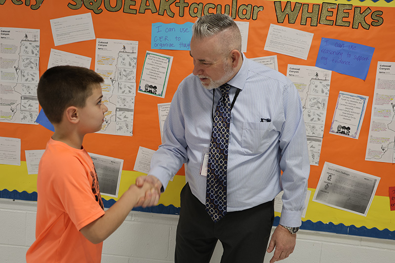 A fourth-grade boy wearing an orange tshirt shakes hands with a man in a shirt and tie.