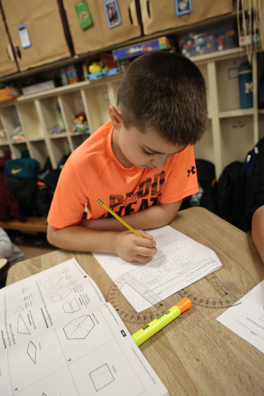 A fourth-grade boy wearing a bright orange shirt does math at his desk. There is a highlighter and a math compass on his desk. 