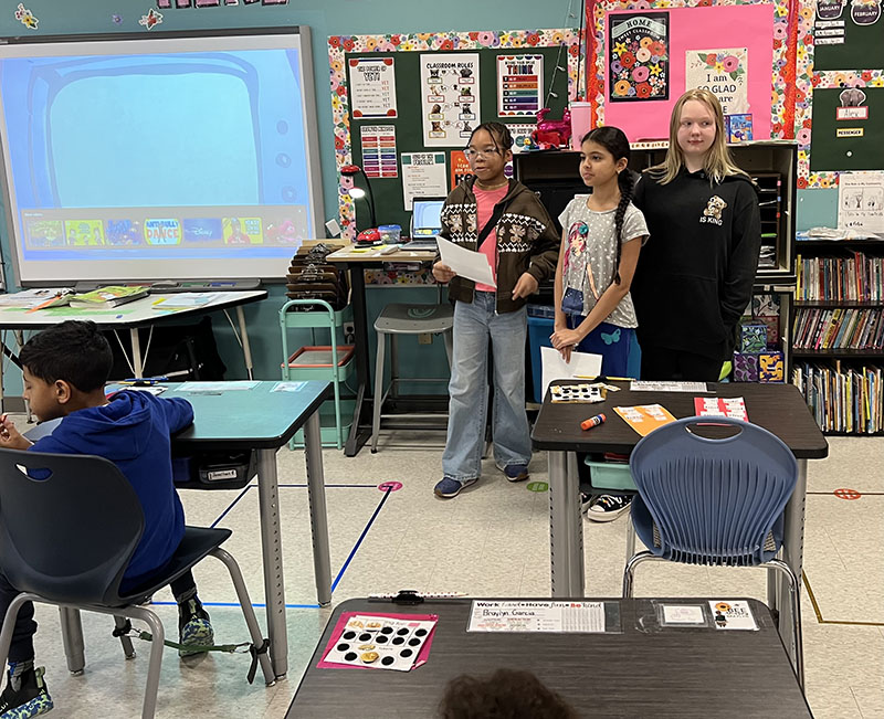 Three fifth-grade girls stand in front of a classroom talking about being an upstander.