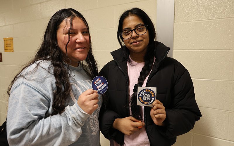 Two high school girls hold up their stickers that say they registered to vote. They are smiling.