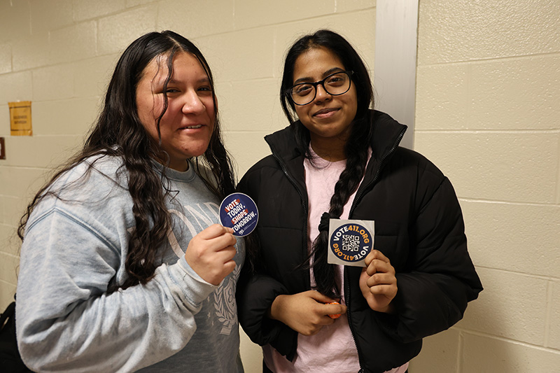 Two high school girls hold up their stickers that say they registered to vote. They are smiling.