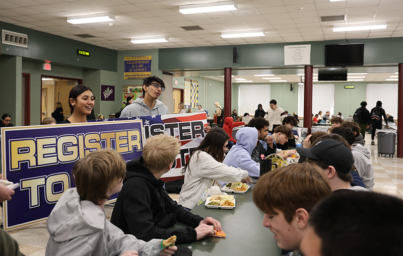 Two high school kids stand near a table of other high school kids. They are holding posters that say Register to Vote.
