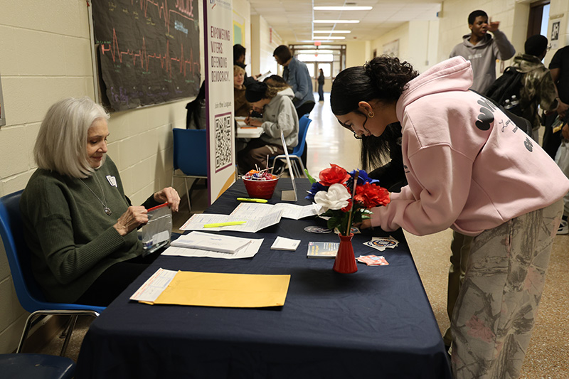 A woman sits on one side of a table and a high school girl leans in to complete a form on the other side of the table.