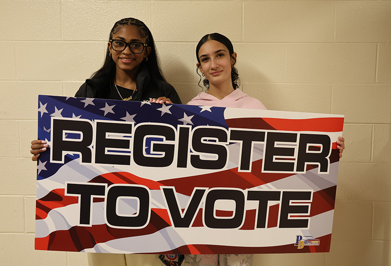 Two young women, both high school students, hold a big sign that says Register to Vote.