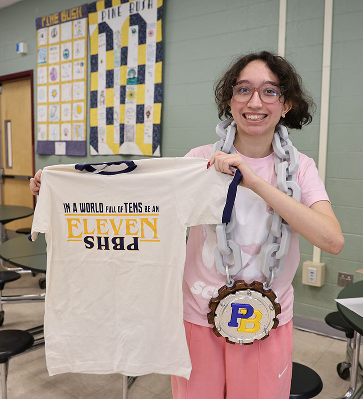 A high school junior smiles as she holds up a white tshirt that says In a world of 10s be an eleven PBHS. She is also wearing a large plastic chain around her neck with a pendant PB.