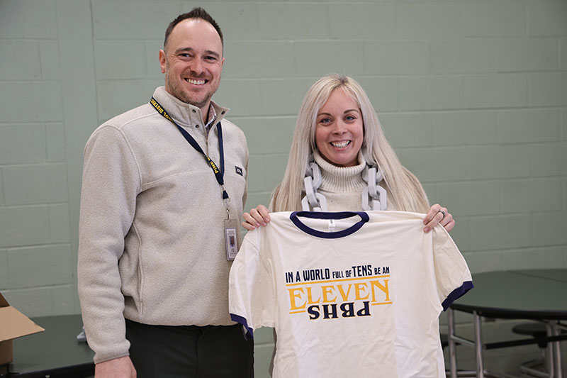 A woman with long blonde hair smiles as she holds up a white tshirt that says In a world of 10s be an eleven PBHS. Next to her is a man also smiling.