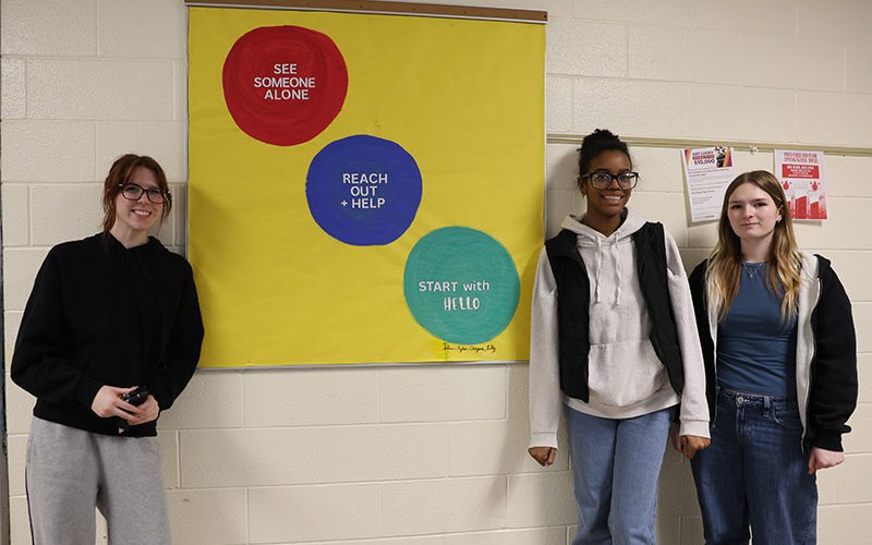 Three high school girls stand next to a bulletin board they just created.