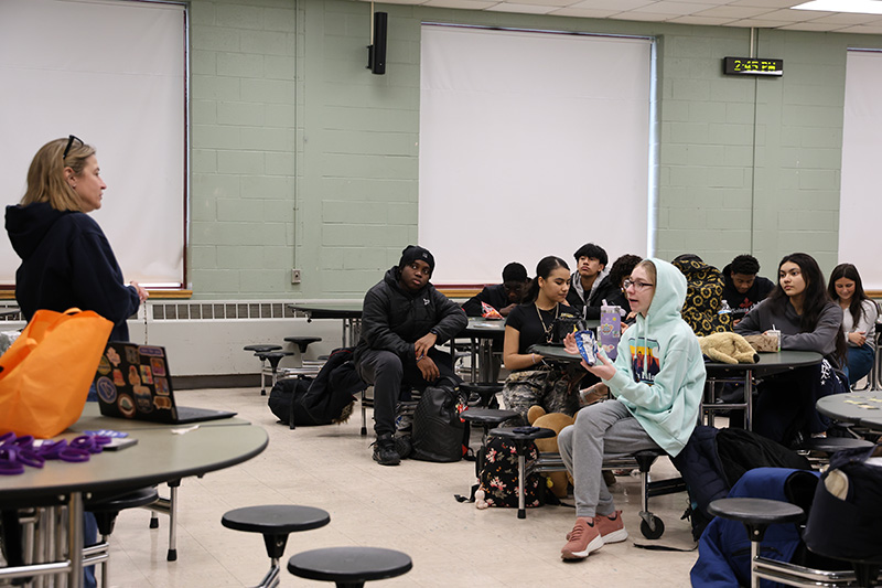 A woman in a blue sweatshirt stands in front of a large group of high schools students who are sitting at tables.
