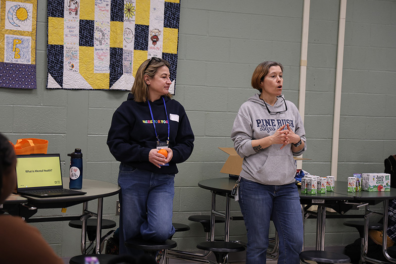 Two women stand in front of a group of high schools students, talking to the kids.