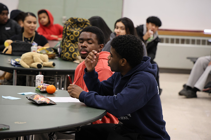 High school kids sit at tables in a cafeteria. One young man is speaking.