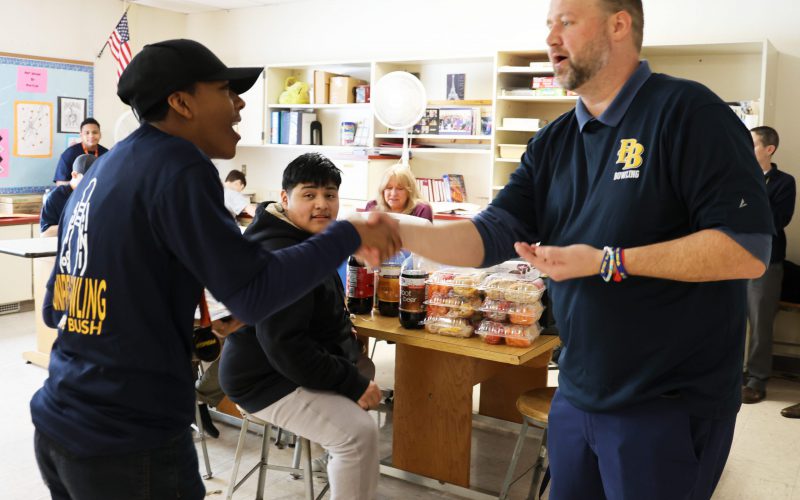 A high school boy wearing a blue baseball cap shakes hands with a man.