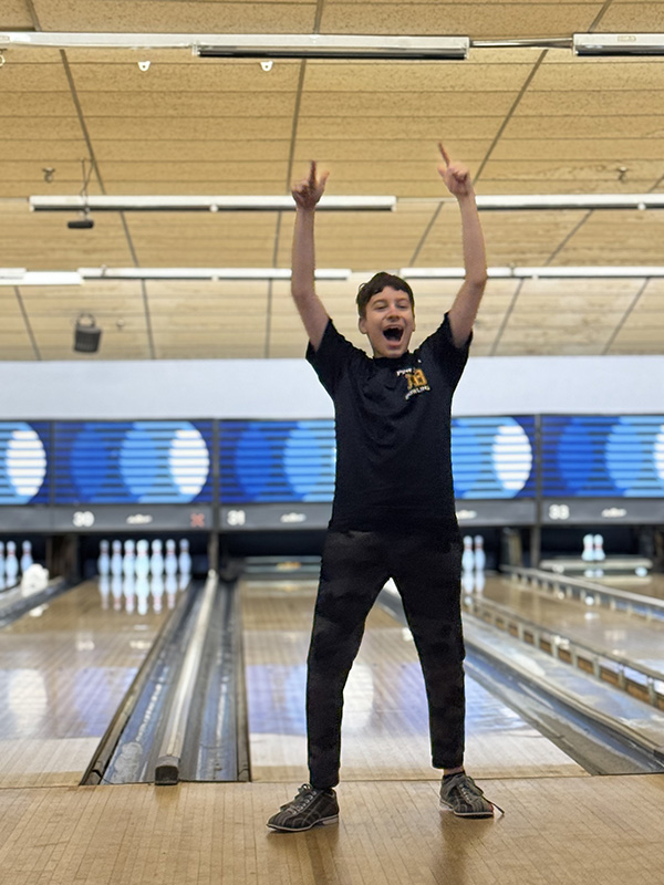 A high school boy holds his arms up in excitement. He is at a bowling lane. 