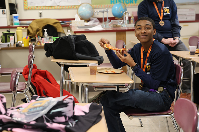 A high school boy smiles as he eats pizza.