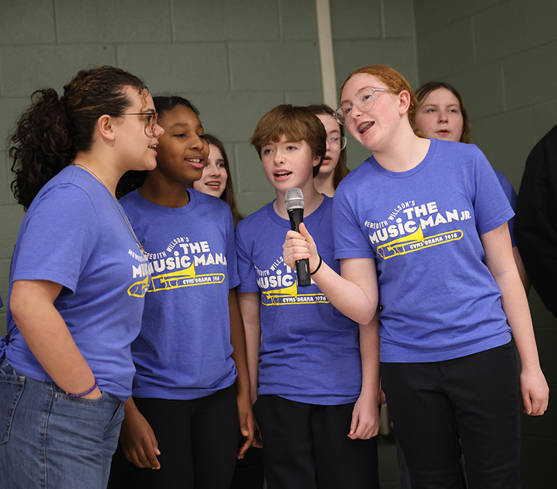 Four middle school students wearing blue shirts that say The Music Man Jr. sing together.