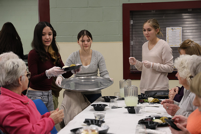 Three high school girls serve dinners to senior citizens. 