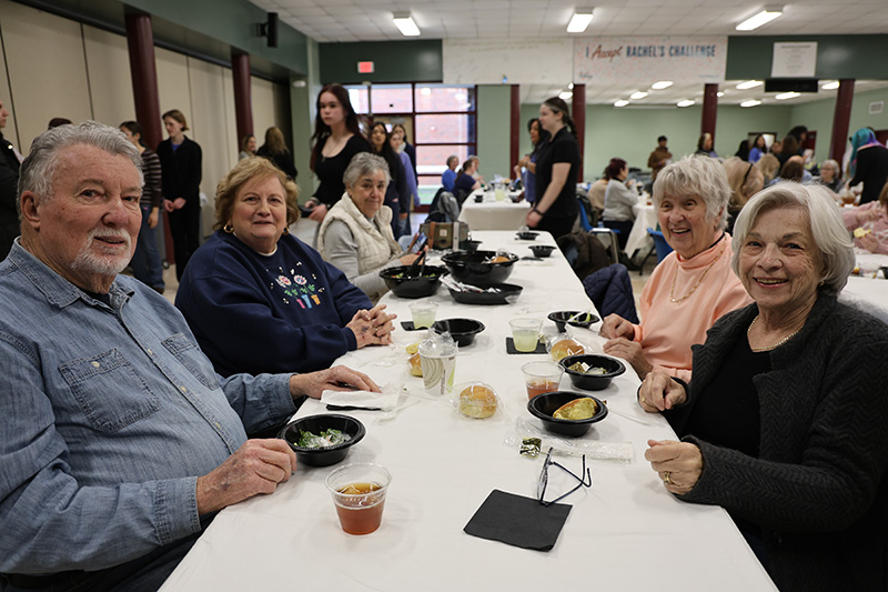 A group of senior citizens sit at a table together waiting to eat. They are smiling.