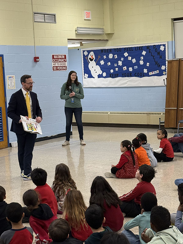 A man stands in front of a group of elementary age kids and reads a book to them.