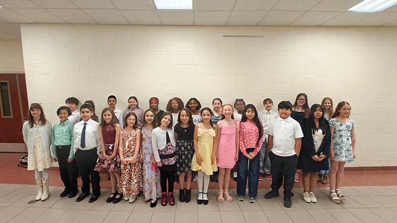 A group of 26 elementary and middle school students stand together in two rows. They are all dressed nicely in dresses and dress shirts and pants.