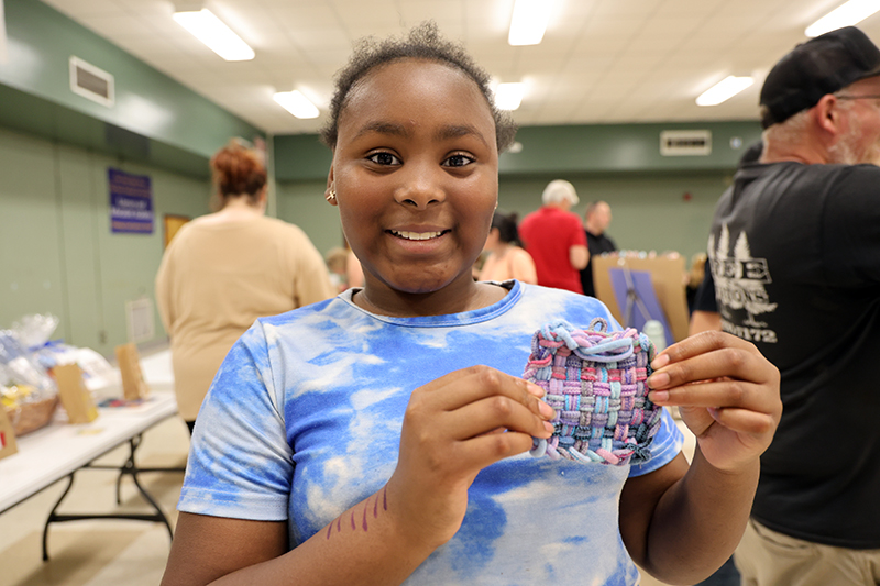 A younger girl in a blue and white tie dye shirt smiles and holds up a pot holder she made.
