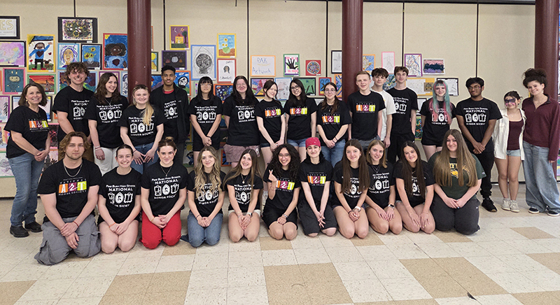 A large group of high school kids and a couple of adults all wearing dark blue tshirts pose in two rows for the picture. They are smiling.