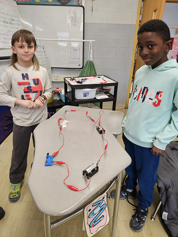 Two fourth-grade students stand at a desk that has lightbulb, and wires.